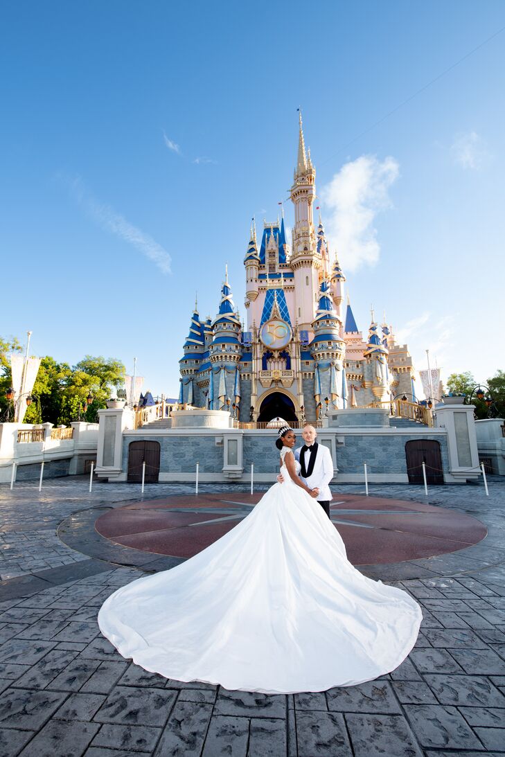 A bride in a princess ballgown with a long train and a crystal headpiece and a groom in a white tuxedo jacket with black trim pose together in front of Cinderella Castle in the Magic Kingdom at the Walt Disney World Resort.