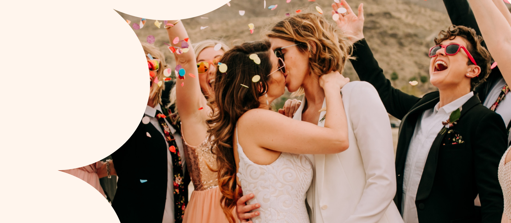 A couple dressed in white kiss surrounded by guests, confetti and mountains.