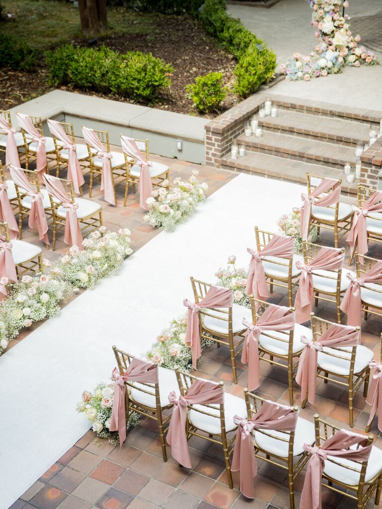 aerial view of outdoor wedding ceremony venue with pink sashes on gold chiavari chairs, floral arch backdrop, white aisle runner and low aisle markers