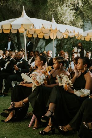 Guests in All Black Outfits, Bridesmaids With Colorful Bouquets Sit at Ceremony Under Bright Umbrellas