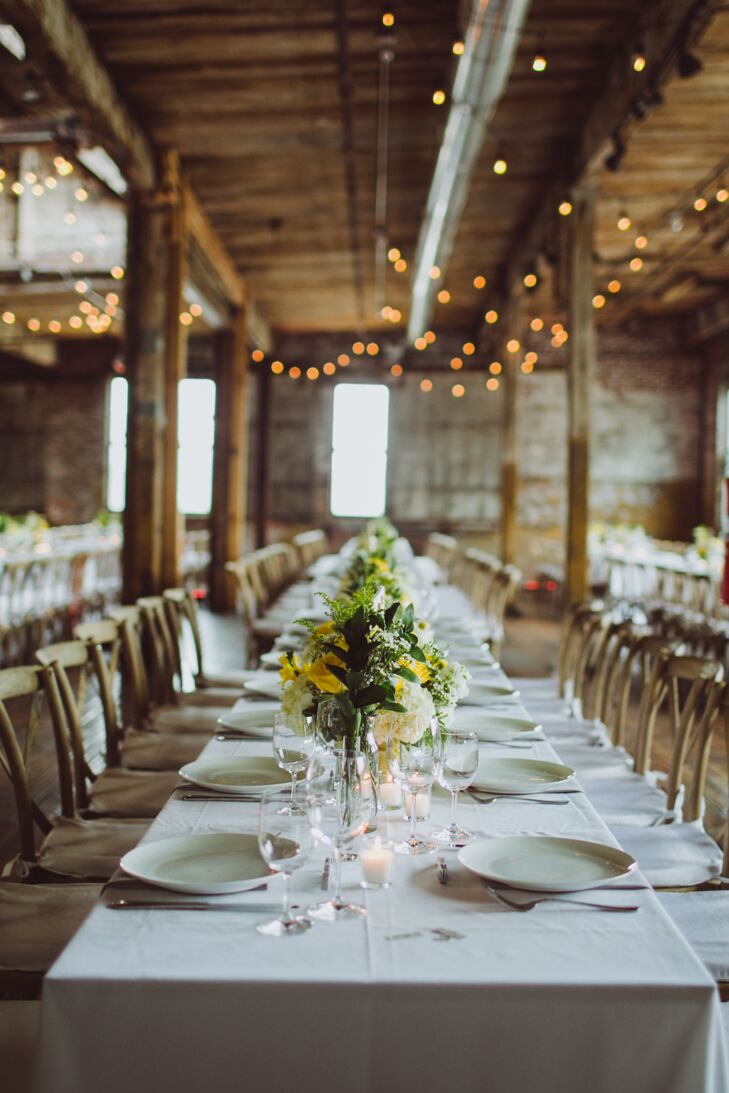For the reception at the Greenpoint Loft, the couple kept the decor simple, letting the venue's features take center stage. Long farm tables draped in white linens were styled alongside cross-back chairs and bundles of brilliant yellow and green blooms. Twinkling bistro lights hung overhead, casting a warm, ambient glow over the expansive loft space.