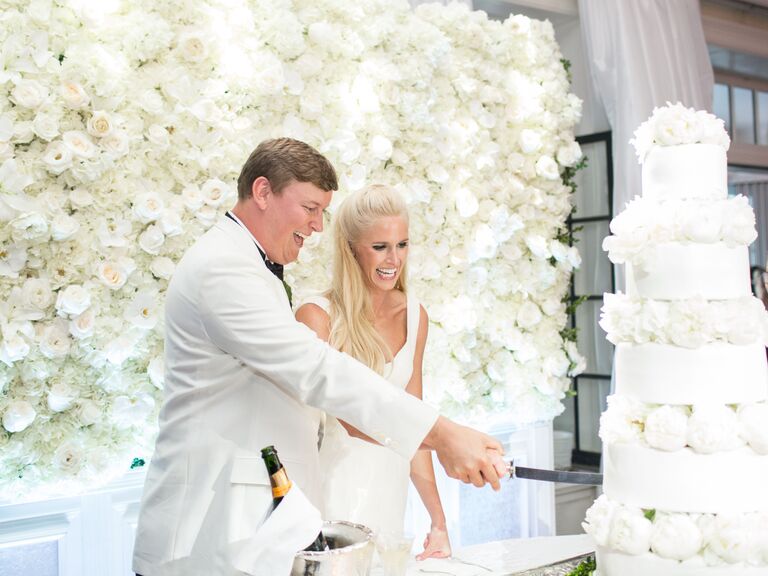 Bride and groom cut their wedding cake with white rose wedding flower wall behind them.
