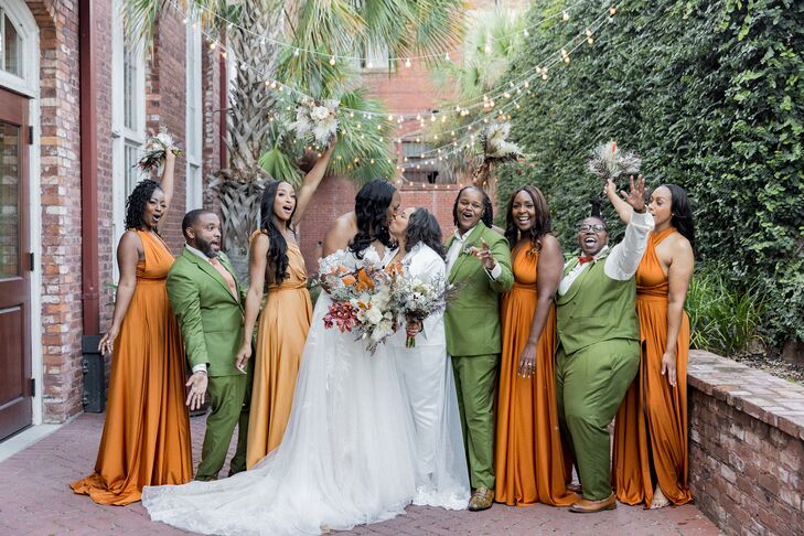 Two brides, one in a white dress and one in a white pantsuit and sheer overskirt, kiss amid their mixed bridal party in burnt orange dresses and hunter green suits with fall-themed bouquets at their bohemian-chic weddings.