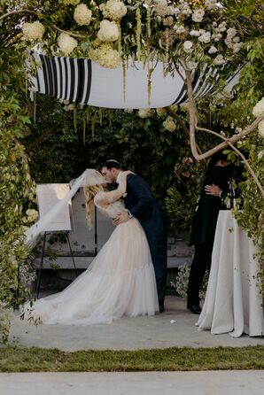 Groom in Navy Tuxedo Dipping Bride in Lace-and-Tulle Gown Back for First Kiss Under Chuppah, Greenery