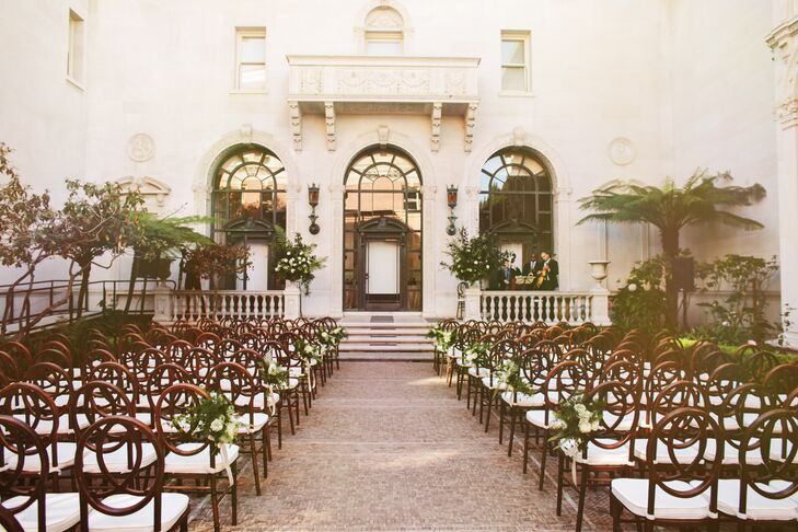 The ceremony was held outside the elegant historic building of the The James Leary Flood Mansion in San Francisco, California. Elegant dark wooden chairs with white cushions were lined up in rows, facing the altar where the couple were  married. Small flower arrangements filled with ivory and green accents were tied to the end of the chairs in the aisle.