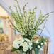 To inject an element of drama into the escort card display, Botanica International Design Studio centered the golden seashell card holders on a trio of hurricane vases overflowing with delphiniums, branches, hydrangeas and lisianthus in bright shades of green and white.