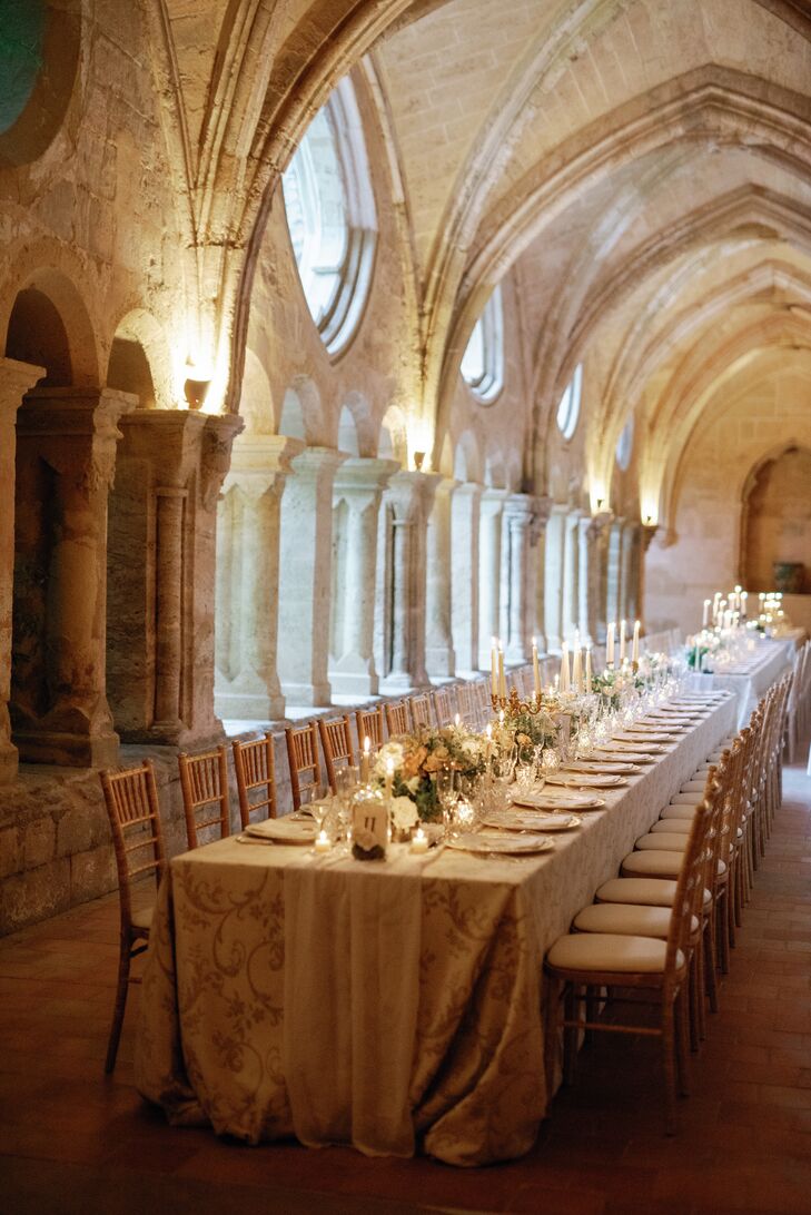 A romantic, low-lit reception space in a historic French Abbey venue with long tables, ornate linens, low and tall candles, low centerpieces and a neutral color palette to accent the gorgeous space.