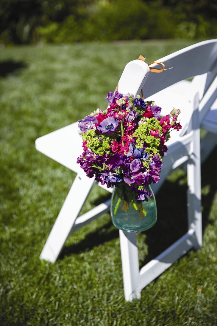 Purple and magenta stock ranunculuses, anemones, tulips, garden roses, and lisianthuses were displayed in clear glass vases and hung on the aisle chairs.