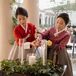 The mother of the bride and the mother of the groom in traditional Korean hanbok dresses (one in red and one in purple) lighting a unity candle surrounded by greenery at the altar during a Christian ceremony.