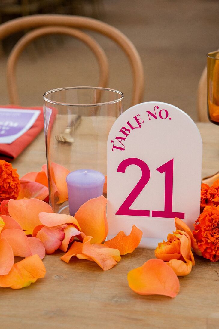 Half-dome table number with hot pink writing at bold fusion wedding with flower petals, a lavender candle and orange flowers on the tablescape.