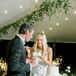 Groom in Classic Tuxedo and Bride With Down Hairstyle Cutting, Feeding White Wedding Cake