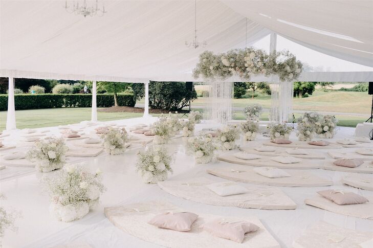White and neutral-toned Sikh wedding ceremony with acrylic aisle, white floral aisle decor, plush floor cushions for guests and a Guru Granth Sahib altar with hanging flowers and crystals.