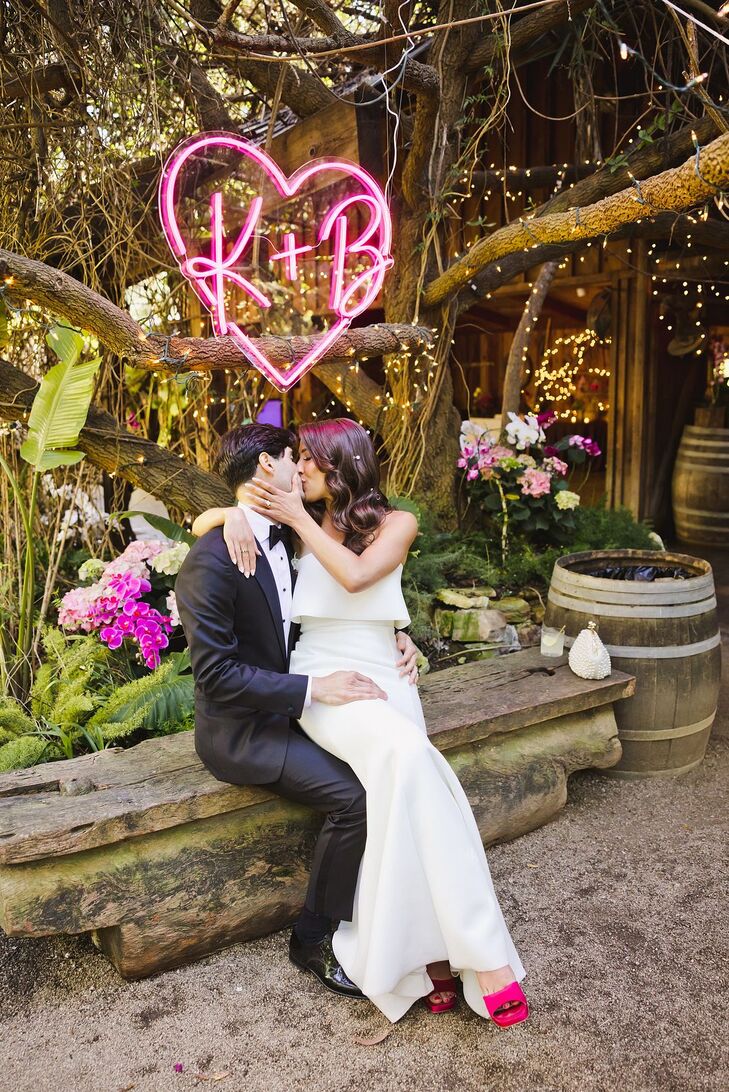 A groom in a navy tuxedo kisses his bride, wearing a sleek fitted gown with a strapless bodice and hot pink shoes underneath a custom retro neon sign with the couple's initials amongst bright-toned flowers in the garden of their wedding venue.