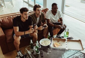 group of three men sitting on a brown leather sofa playing video games and eating snacks and beer