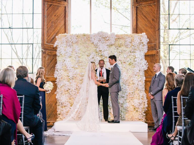 Bride and groom stand in front of a white and pale yellow flower wedding wall during a ceremony.