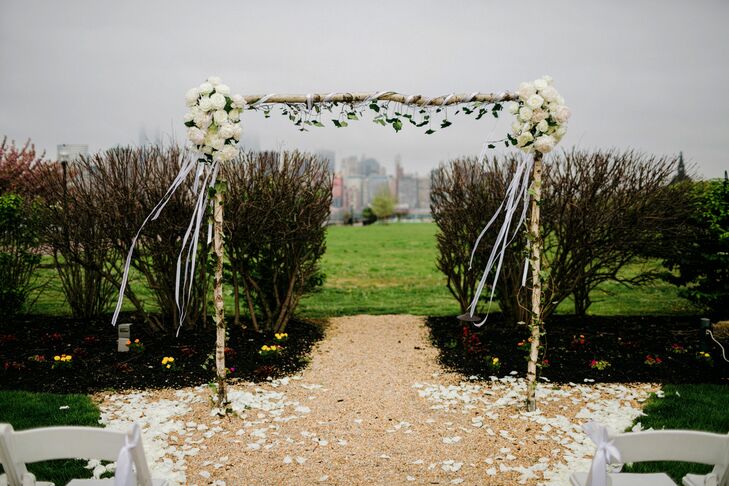 Keeping with their natural style, Cory Davenport designed a chic birchwood wedding arch for the outdoor fete at Liberty House Restaurant in Jersey City, New Jersey. Ivy wrapped around each post in between strands of white ribbon. Bringing the two elements together, their florist added bunches of white roses to each corner. Cascading white rose petals also covered the ground for a classic touch.