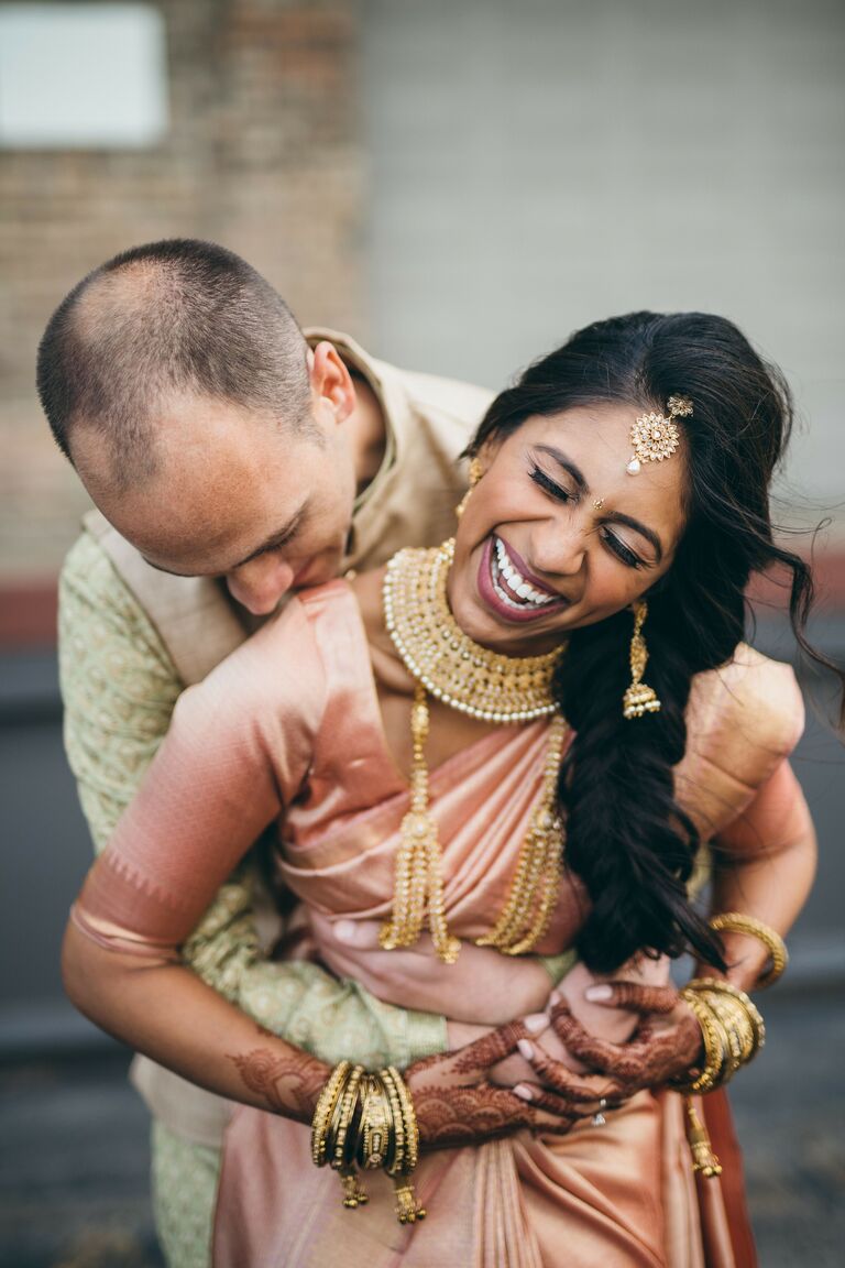 candid photo of couple laughing and hugging while wearing traditional Indian wedding attire in 2022 wedding colors sage green and light peach
