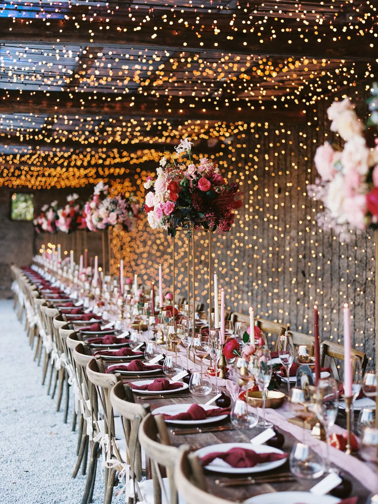 A fairytale table setting of maroon napkins, pink and maroon tapers, and twinkling string lights.