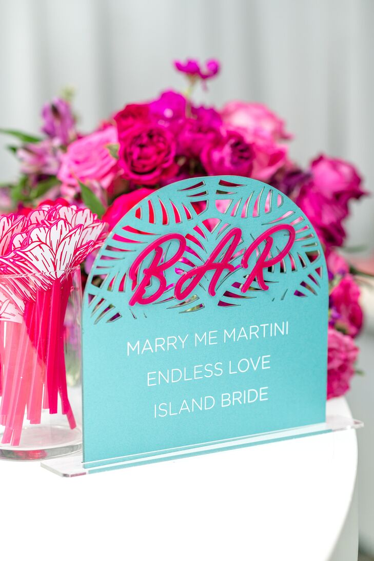 A bright blue, patterned bar sign with pink lettering featuring three signature cocktails for a bridal shower: a Marry Me Martini, an Endless Love and an Island Bride with custom, pink, floral stirrers.