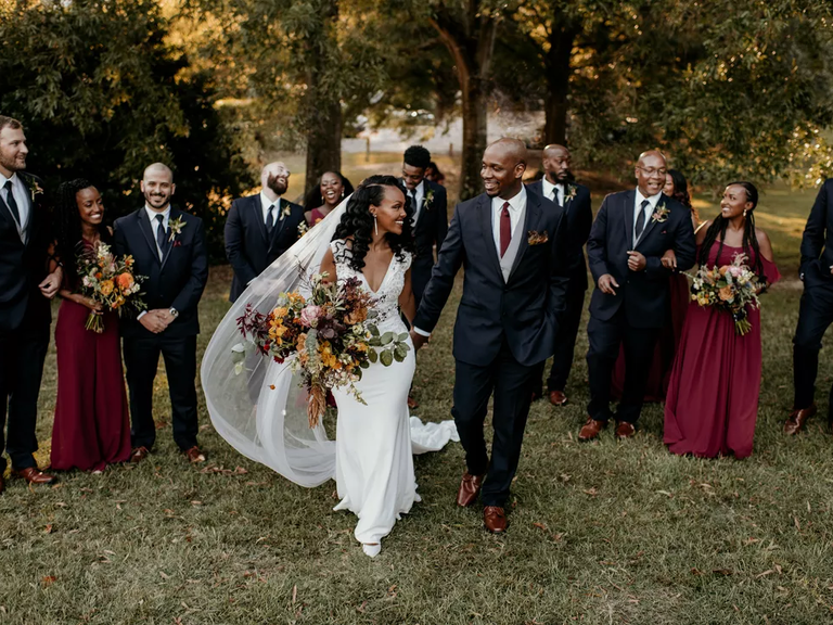 A bride and groom walk towards the camera, surrounded by their beaming wedding party.
