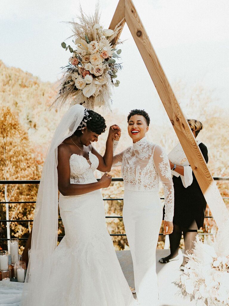 Brides holding hands in front of angular floral ceremony arch at outdoor summer wedding