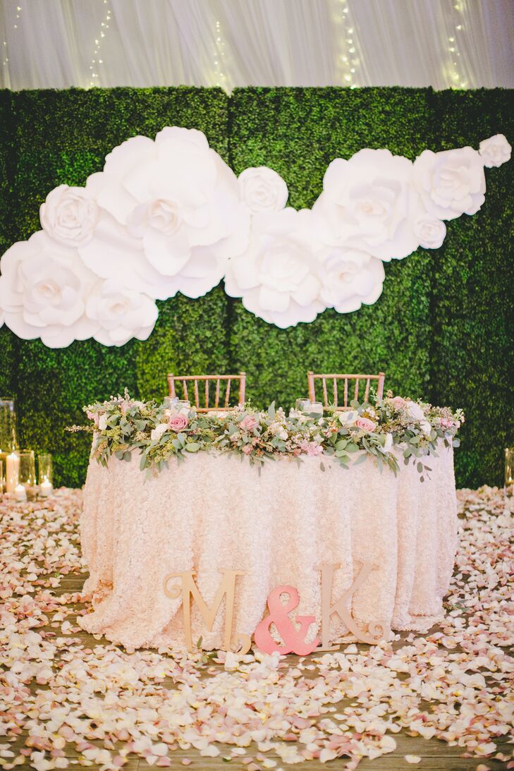 Marisa and Kyle sat at their sweetheart table draped with a lush garland, filled with romantic blooms and greenery. Pink cherry blossom petals surrounded the table, positioned in front of a large gardenia wall decoration on a trimmed hedge backdrop.