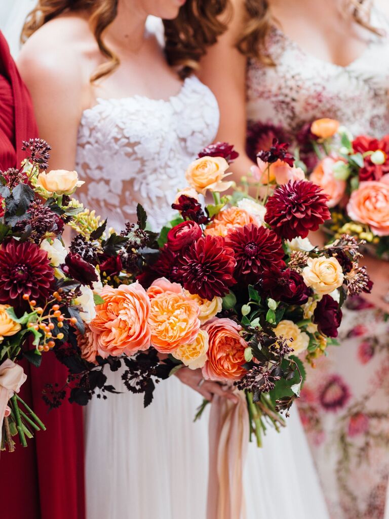Bride and bridesmaids hold vibrant bouquets of dahlias and roses in red and orange hues. 