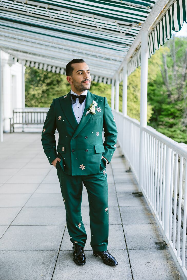 A groom in an emerald-green tuxedo jacket and slacks with a white shirt and a black bow tie with jewels all over the tuxedo jacket posing before his wedding at The Boathouse in New York City.