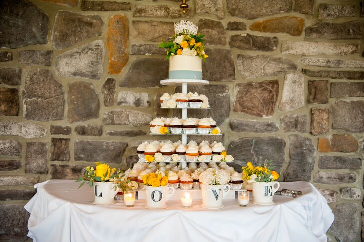 The tall stand displayed a variety of cupcakes, with the white wedding cake with a light blue band wrapped around the bottom of the perched top. The cake had yellow and ivory blooms that matched the flowers inside the vintage-styled mugs spelling out "love."