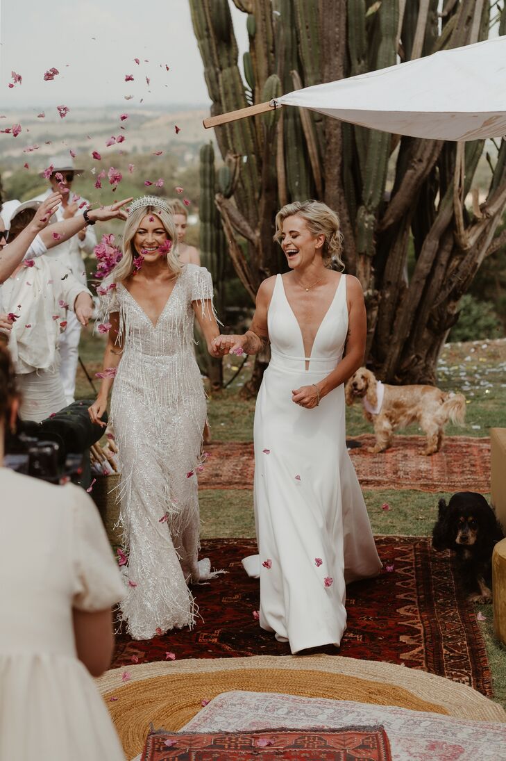 A bride in a fully beaded, couture wedding gown with beaded tassels and a diamond tiara walks up the aisle with oriental rugs in processional with her bride, dressed in a different, sleek wedding gown with a plunging neckline and a sophisticated updo in a desert setting with cacti as guests throw flower petals.