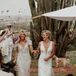 A bride in a fully beaded, couture wedding gown with beaded tassels and a diamond tiara walks up the aisle with oriental rugs in processional with her bride, dressed in a different, sleek wedding gown with a plunging neckline and a sophisticated updo in a desert setting with cacti as guests throw flower petals.