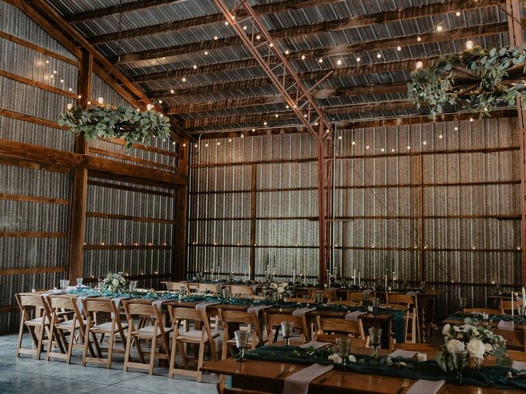 barn wedding reception venue with greenery chandeliers and bistro string lights hanging from vaulted wooden rafters