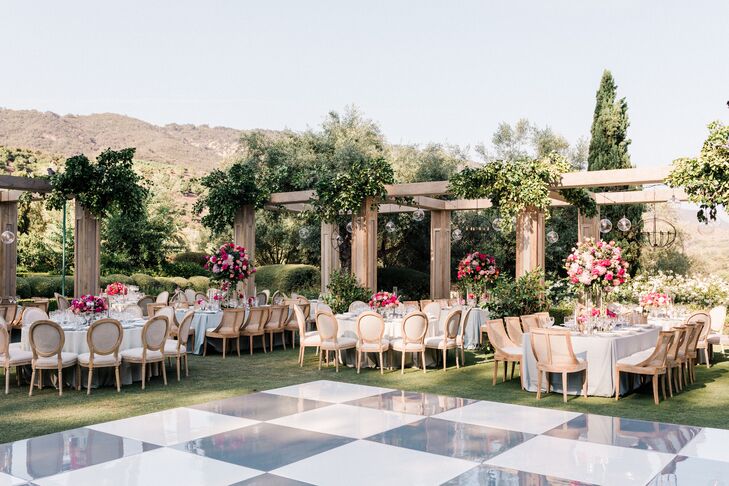 A couple's elegant garden reception space at an estate in Santa Barbara, featuring custom pergolas, hanging greenery, round tables, tall jewel tone floral centerpieces and a custom dance floor.