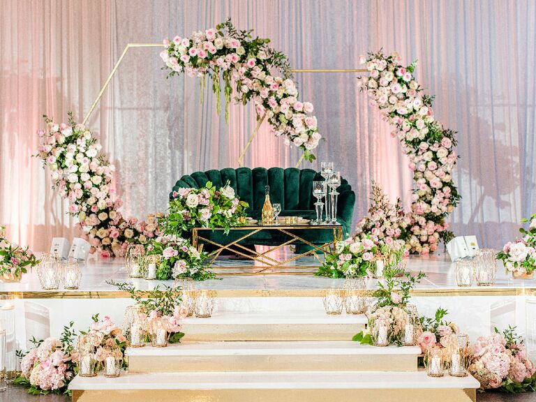 Glam sweetheart table with green velvet couch and gold floral arches