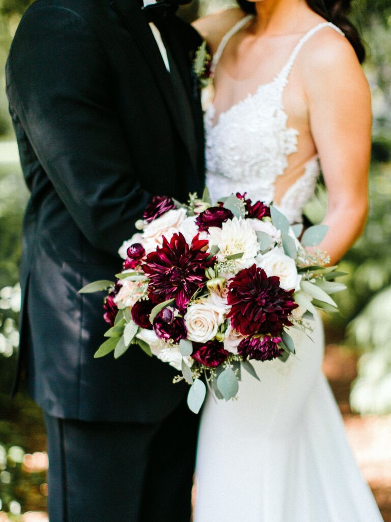 Couple embrace while holding a burgundy and white bridal bouquet. 