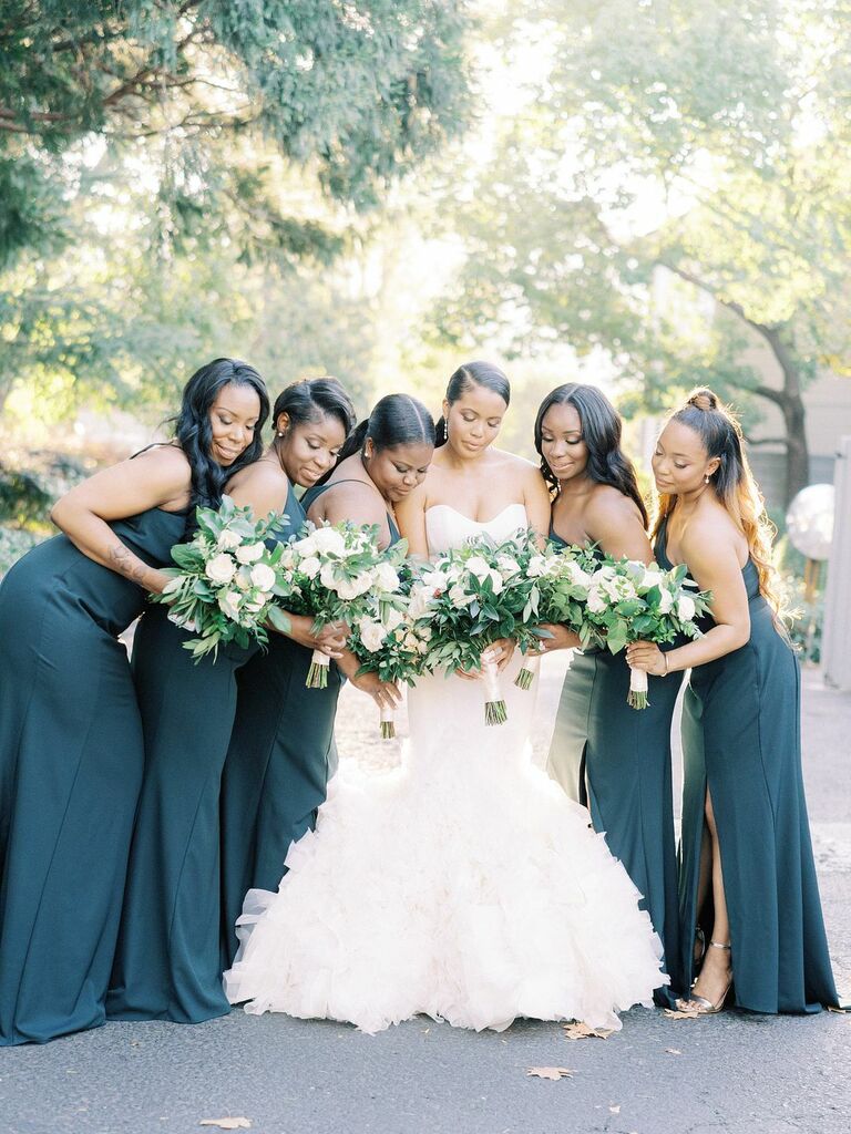 bride wearing ruffled mermaid wedding gown stands outside next to bridesmaids in floor-length matching navy blue gowns holding white and greenery bouquets