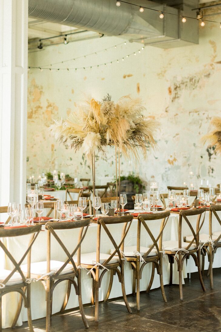 A neutral-toned, bohemian-chic, fall reception design with long tables, burnt orange napkins, string lights and tall pampas grass centerpieces at a loft venue.