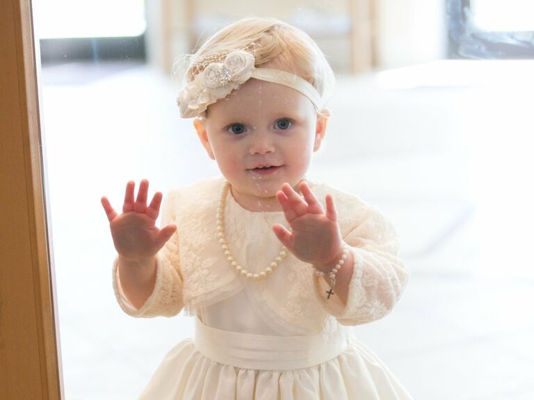 Flower girl looks in a window while wearing a white headband in her hair. 
