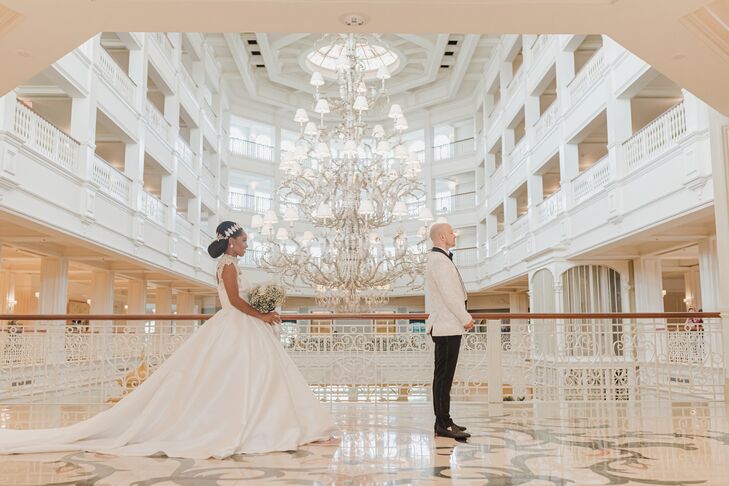 A whimsical bride in a princess-like ballgown with crystal neckline details, a crystal headpiece and a long train walks to greet her groom, in a white tuxedo jacket with black trim, for their first look in a Victorian-inspired hotel at the Walt Disney World Resort.