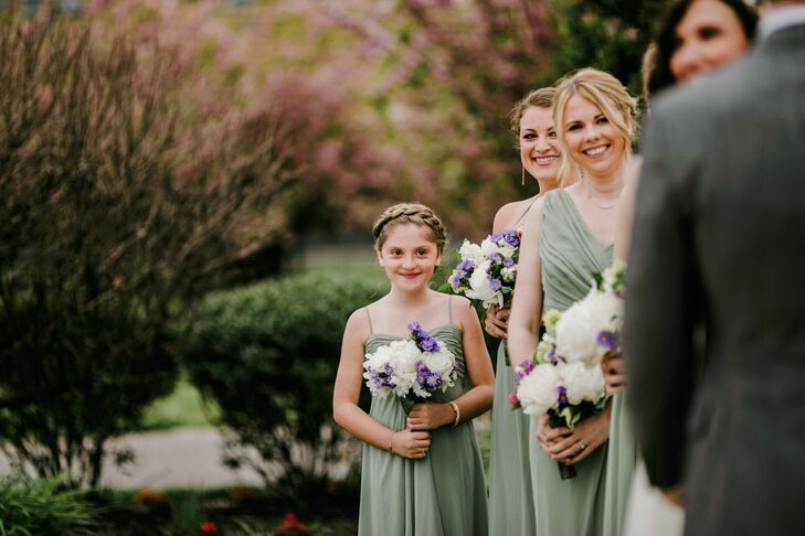 Janel and Charlie’s junior bridesmaid stood out among the group with her own personal style. She donned a classic sage green chiffon dress from Bella Bridesmaids with thin straps and ruching along the bodice, similar to the other women. But she finished it with a bohemian braided updo that wrapped around her like a headband. (How cool is that?) She also carried a slightly smaller bouquet of white roses and purple delphiniums for a welcome pop of color.