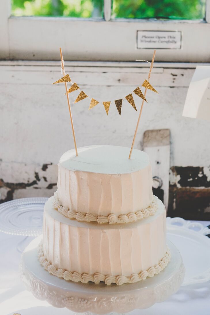 The two-tier ivory wedding cake was covered in buttercream and had vanilla-flavored cake. The bottom of each tier was bordered with thick texture, and the top was accented with a DIY gold flag topper.