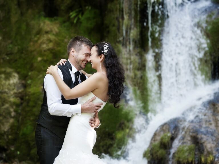 Smiling newlyweds embrace in front of a tropical waterfall. 