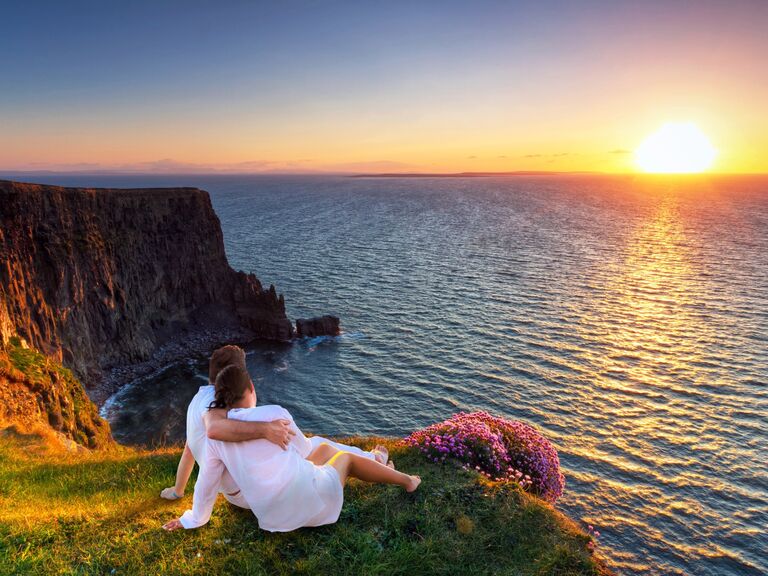  Couple embraces on a cliff edge while watching a sunset over the ocean. 
