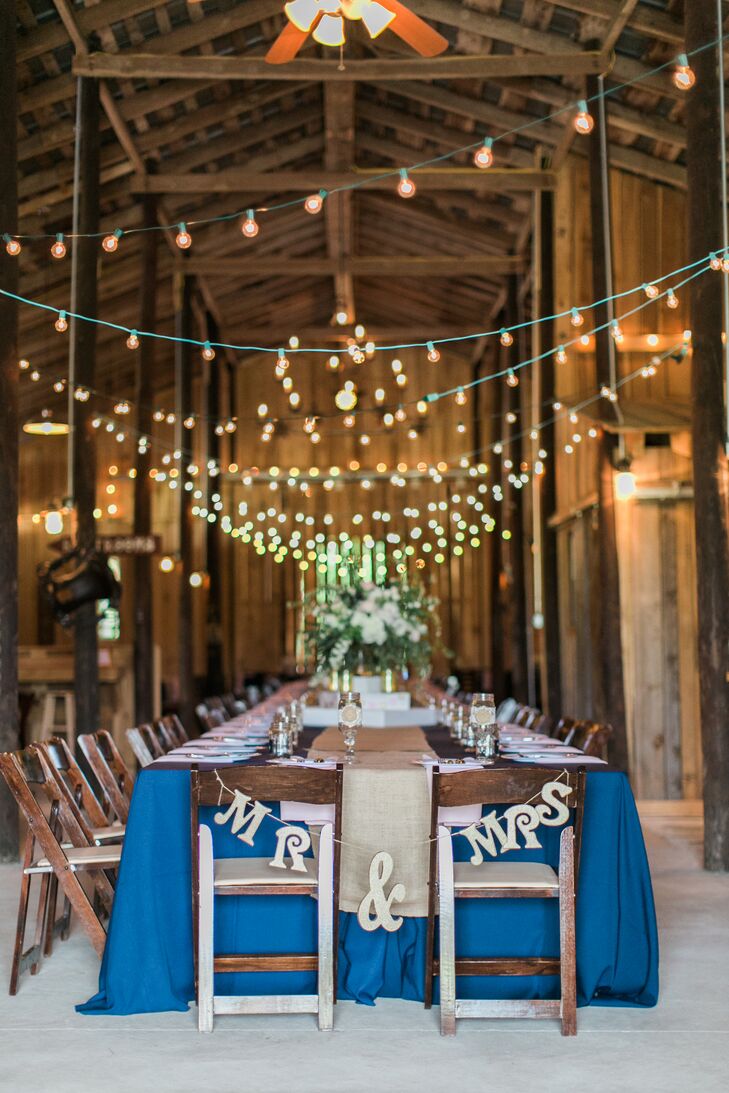 The barn reception featured a long farmhouse dining table that was covered in navy linens, a burlap table runner and blush napkins. String lights hung above the table and the wooden folding chairs. Lauren and Daniel's seats were decorated in a gold "Mr & Mrs" sign.