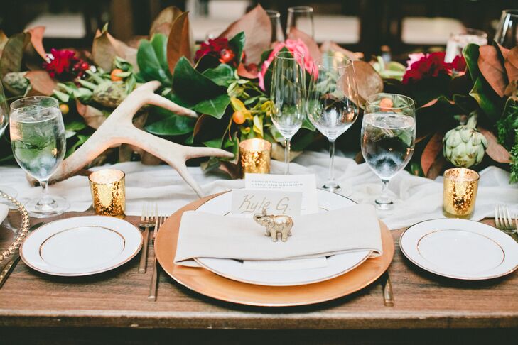 A gold elephant decorated each setting on the wooden dining tables, positioned near the white and gold escort card. White and gold plates contributed to the overall color scheme of the elegant dining display.