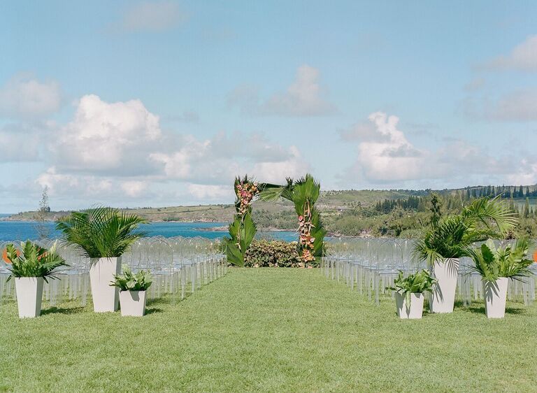 Waterfront wedding ceremony with ghost chairs and tropical leaf arch