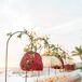 A welcome dinner table with a pink floral patterned linen, low pastel centerpieces and arched, hanging lampposts with red and champagne-colored chandeliers on a hotel balcony overlooking the ocean.