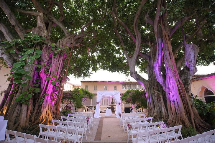 While some couples use minimal decor at The Addison's 100-year-old Banyan tree-lined ceremony space, Irina and Richard added a fun twist. Purple uplighting was set against the trees. This complemented the couple's all-white decor as well as the purple floral accents along the aisle and their wedding arch accent.
