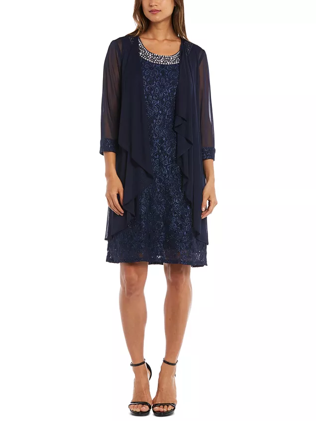 A model wears a petite navy blue wedding guest dress that ends at the knee and features a bejeweled neckline.