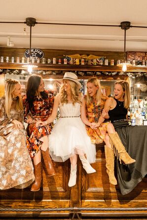 Bride in Short White Dress, Cowboy Boots and Hat With Ladies Sitting on Western Bar